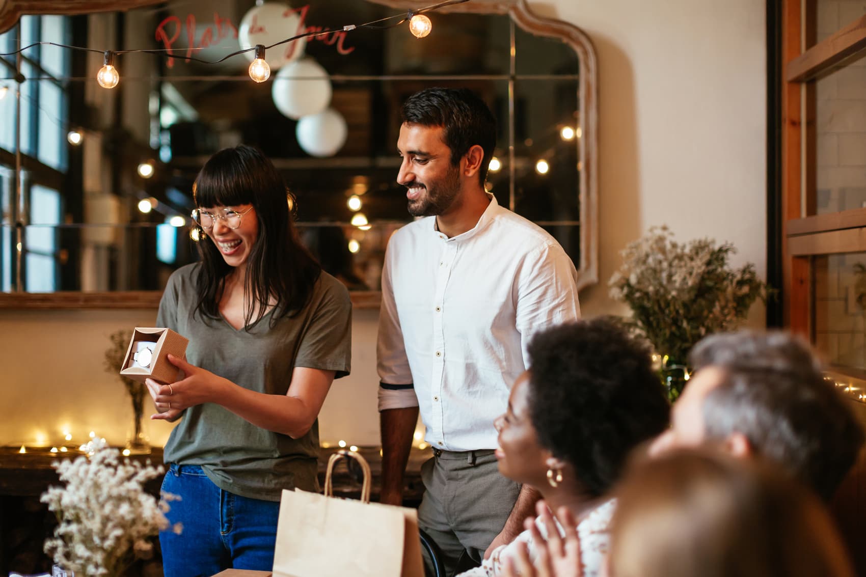 A group of friends gather at a restaurant to celebrate a birthday. A woman stands to show the gift she has just opened — a watch.