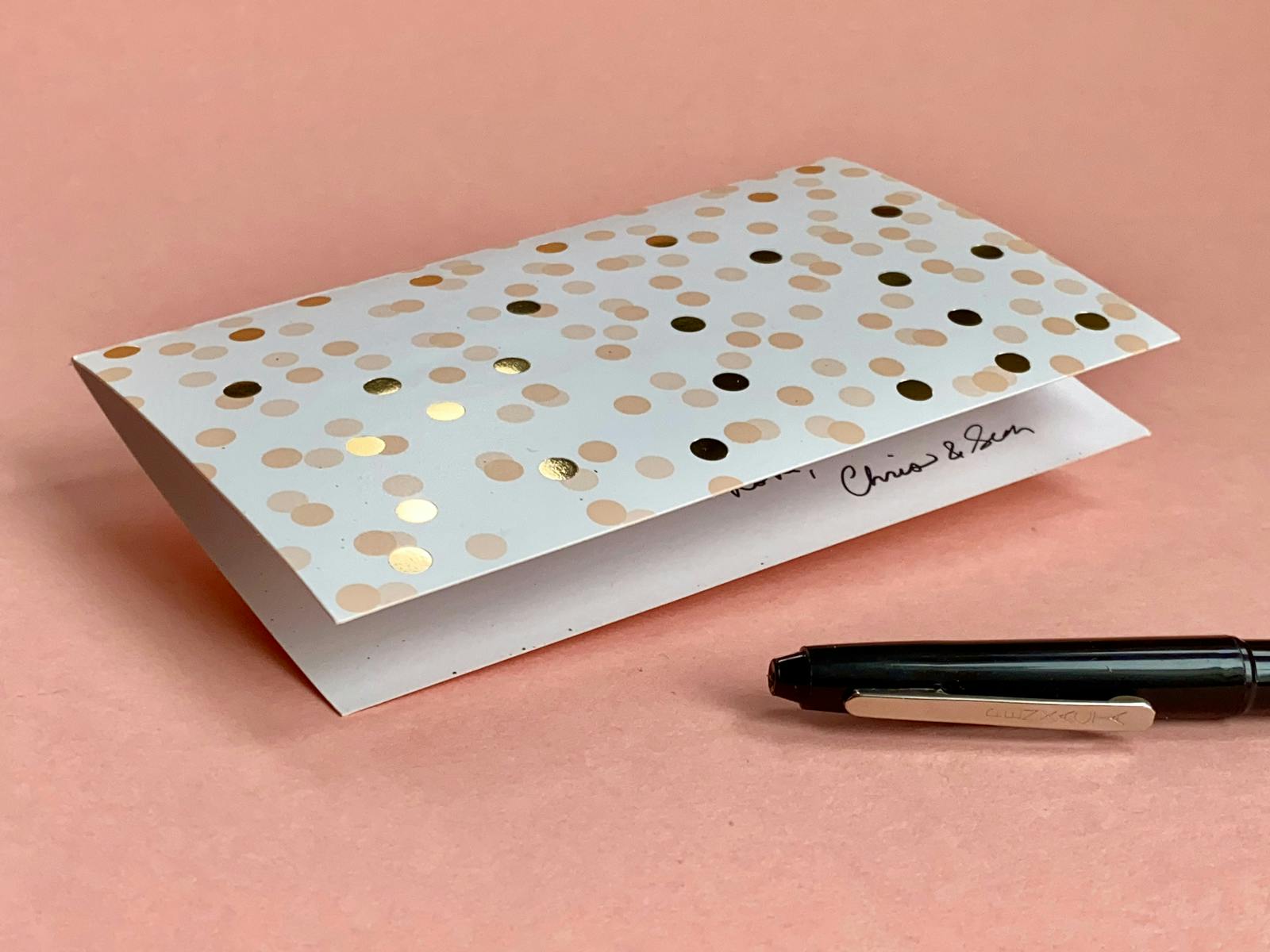 A white birthday card with gold foil dots on a rose pink background. Beside it, a black pen. This is a card our friends sent us that we’ve saved because it was thoughtfully written.