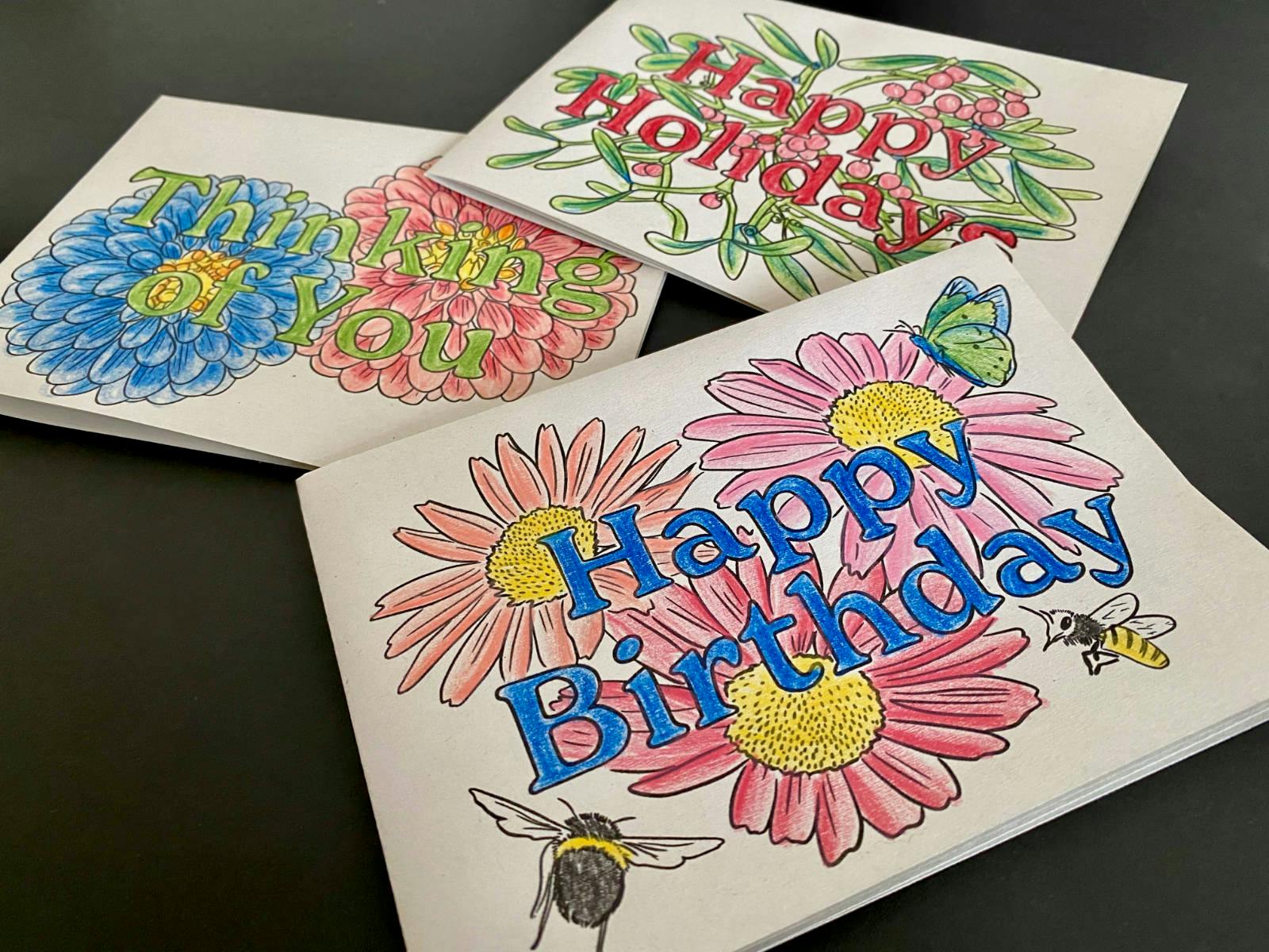 Three handmade cards lay against a black background — one that says “Happy Birthday” one that says “Thinking of You” and the other that says “Happy Holidays”. They have been printed at home and colored in using pencil crayons. You can make these yourself by scrolling to the bottom of the post and downloading the templates for free.