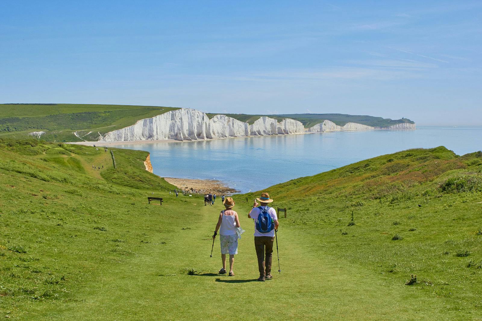 An older couple is walking down a grassy path, facing the white cliffs of the Seven Sisters in Southern England. They are both wearing straw hats. It’s a clear and sunny day.