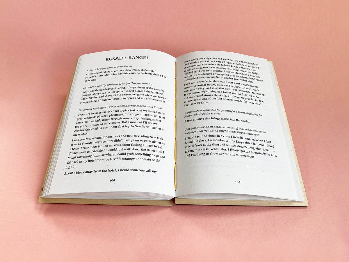 A Fondfolio book with a maple wood cover lays open against a rose pink background. The contributor’s name is printed in uppercase serif type at the top of the left page and their words start right below it and continue onto the right page.